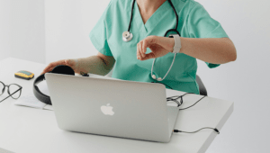 Femme médecin professionnel de santé libéral regardant sa montre connectée devant son ordinateur portable dans son cabinet médical.