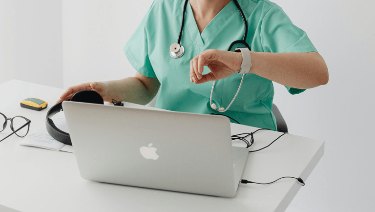 Femme médecin professionnel de santé libéral regardant sa montre connectée devant son ordinateur portable dans son cabinet médical.