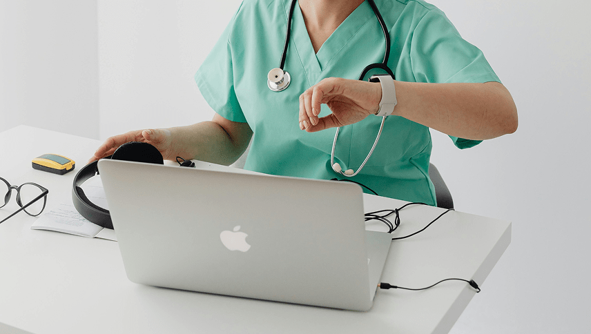 Femme médecin professionnel de santé libéral regardant sa montre connectée devant son ordinateur portable dans son cabinet médical.